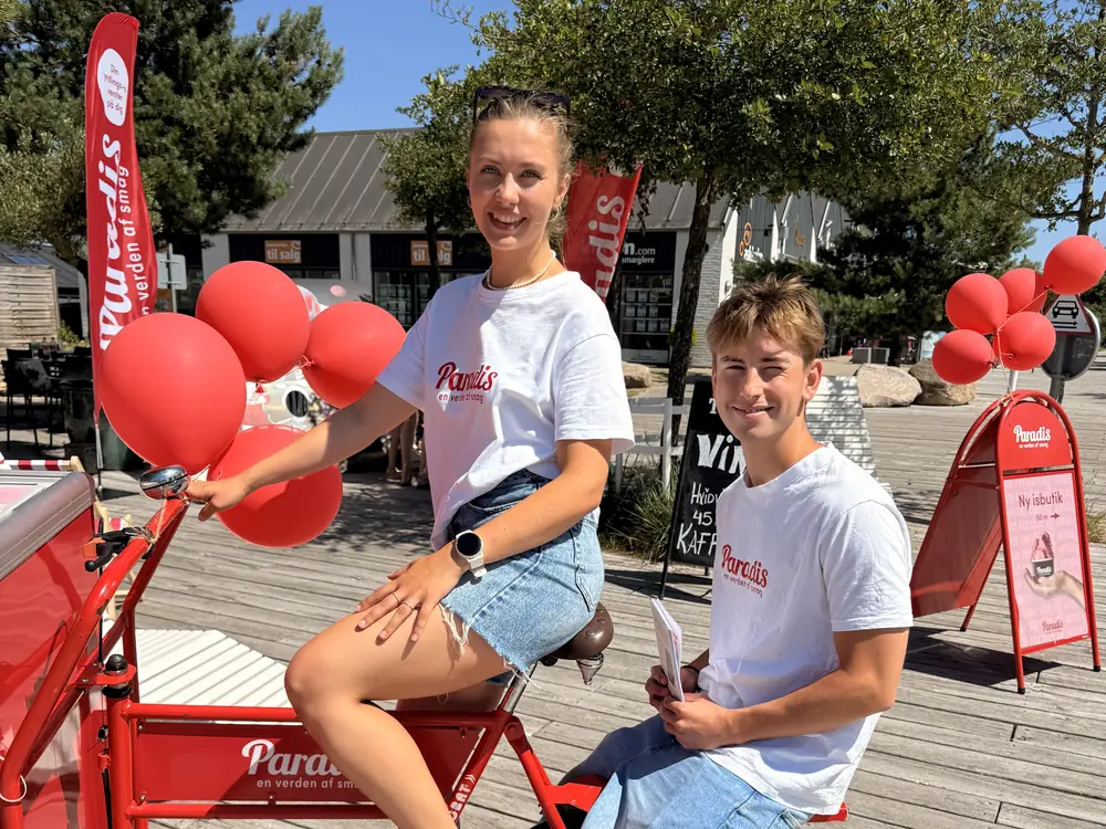 To smilende Paradis-medarbejdere med røde balloner og en rød cykel med isbox